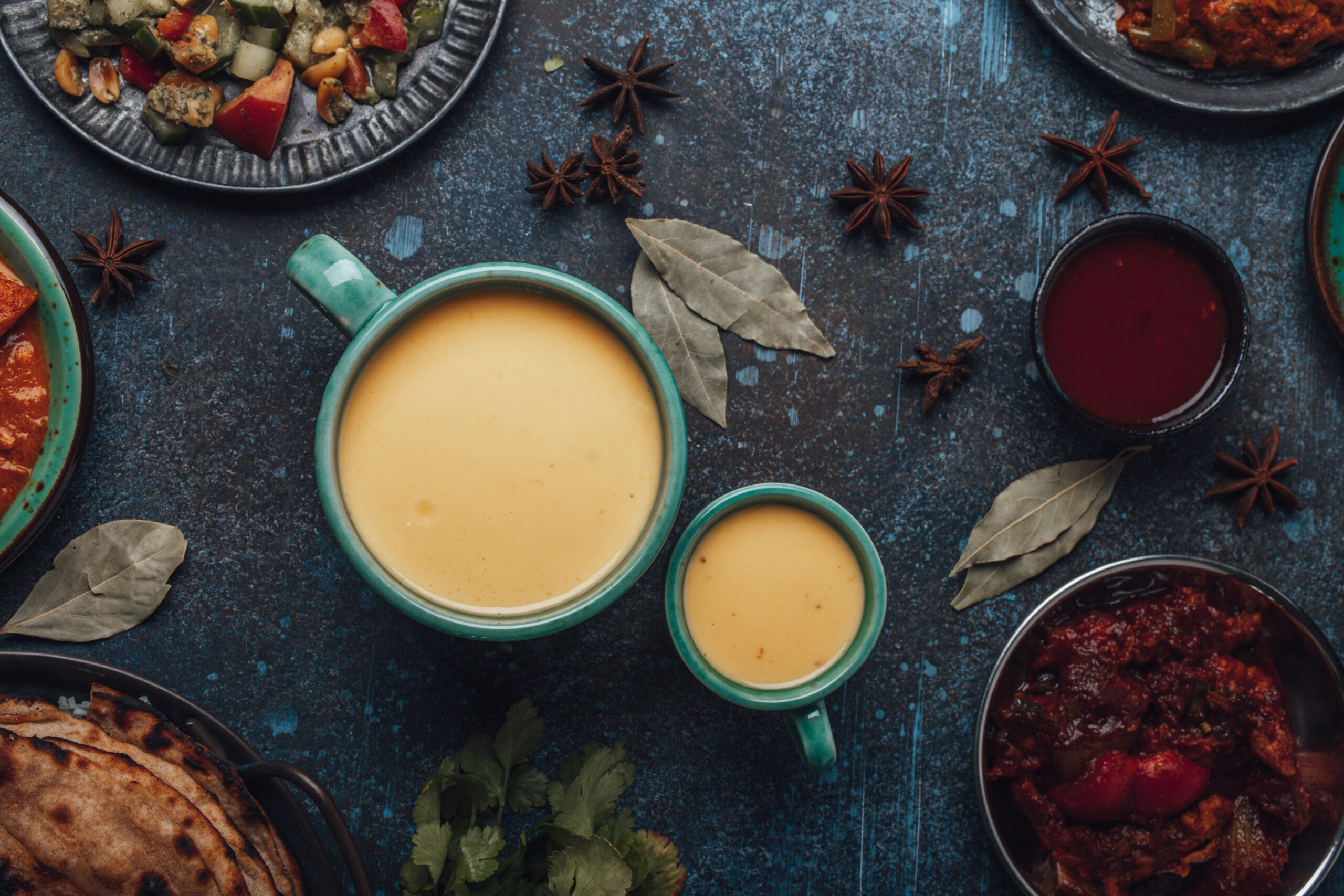 Healthy Indian Ayurveda drink mango lassi in two cups on rustic concrete table with other Indian food from above, yellow blended beverage made of mango fruit, yoghurt or milk curd and spices